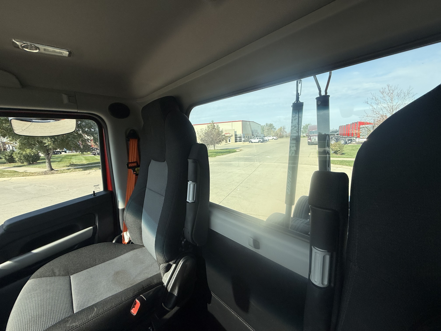 Cab interior of a 2023 Kenworth T680, driver seat and passenger seat, windows visible with road and trucks outside.