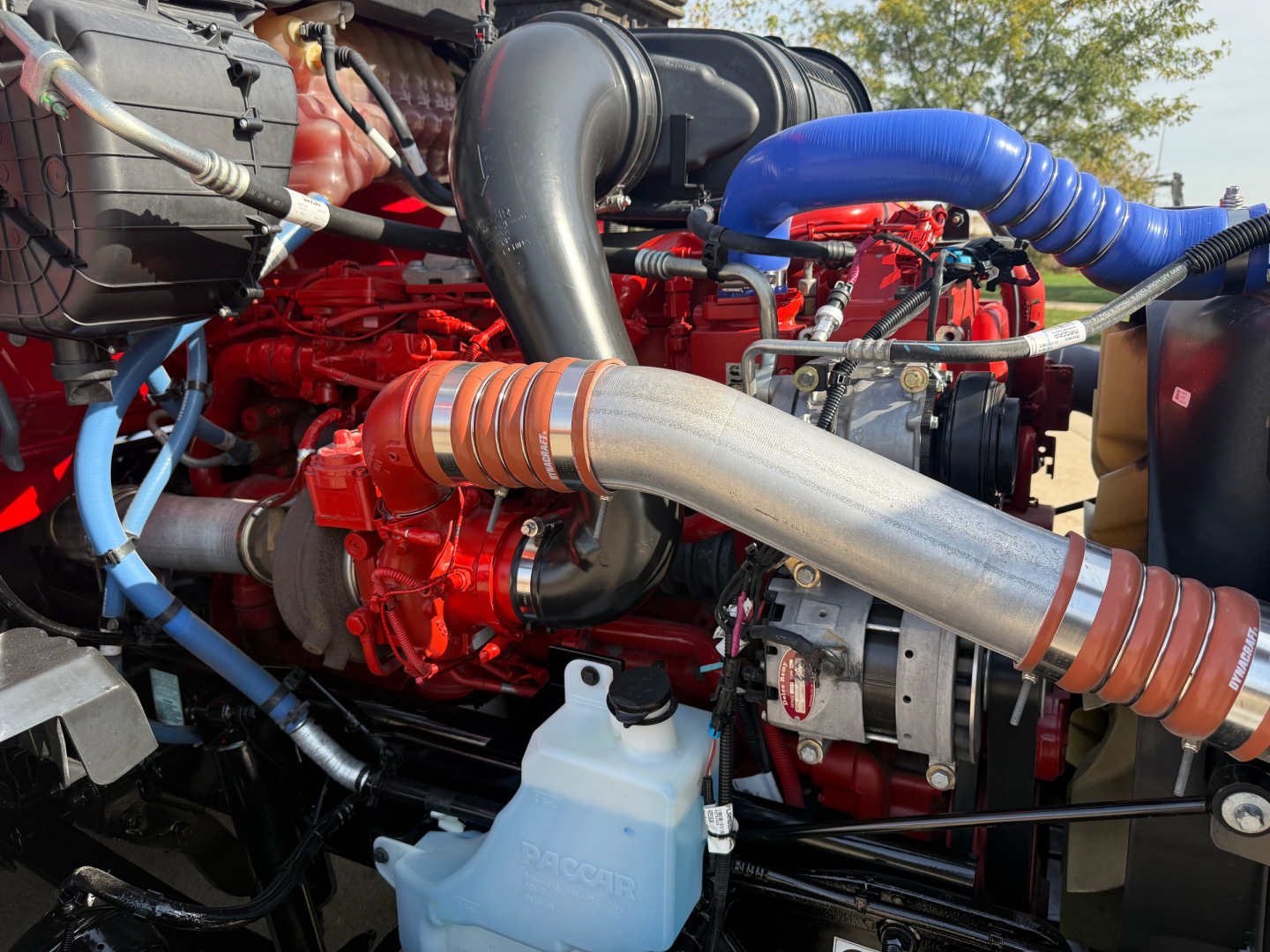 Kenworth T680 engine bay showing large intercooler piping, red engine block, blue hoses, black intake, coolant reservoir with Pa… visible text and components