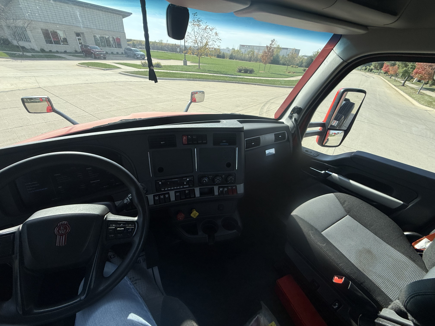 Truck interior view of a 2023 Kenworth T680 cab showing dashboard controls, steering wheel with KW logo, driver and passenger seats, side mirror, and windshield.