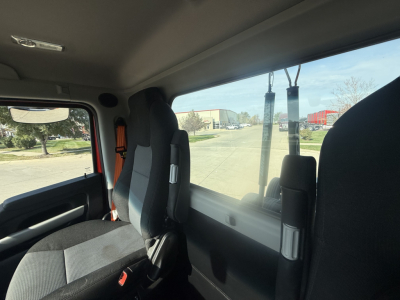 Cab interior of a 2023 Kenworth T680, driver seat and passenger seat, windows visible with road and trucks outside.