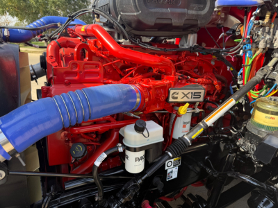 Kenworth T680 engine bay showing a bright red engine with blue hose, labeled 2X15, with white PAS reservoir and various wiring.