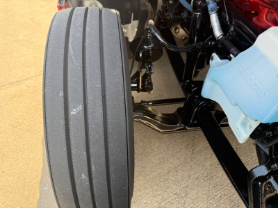 Kenworth T680 front wheel and engine bay area showing large steer tire and coolant reservoir cap; blue coolant reservoir in view; part of chassis and hydraulic lines visible.