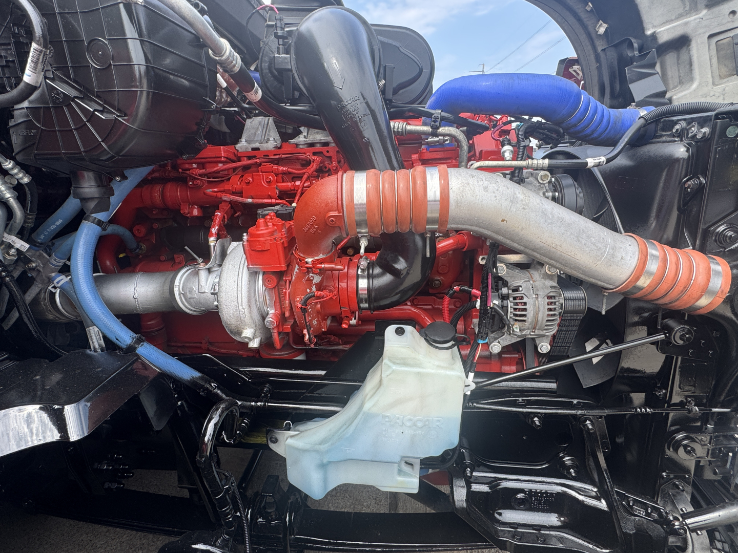 Kenworth T680 engine bay showing red engine, large intake piping, coolant reservoir, and blue hoses.