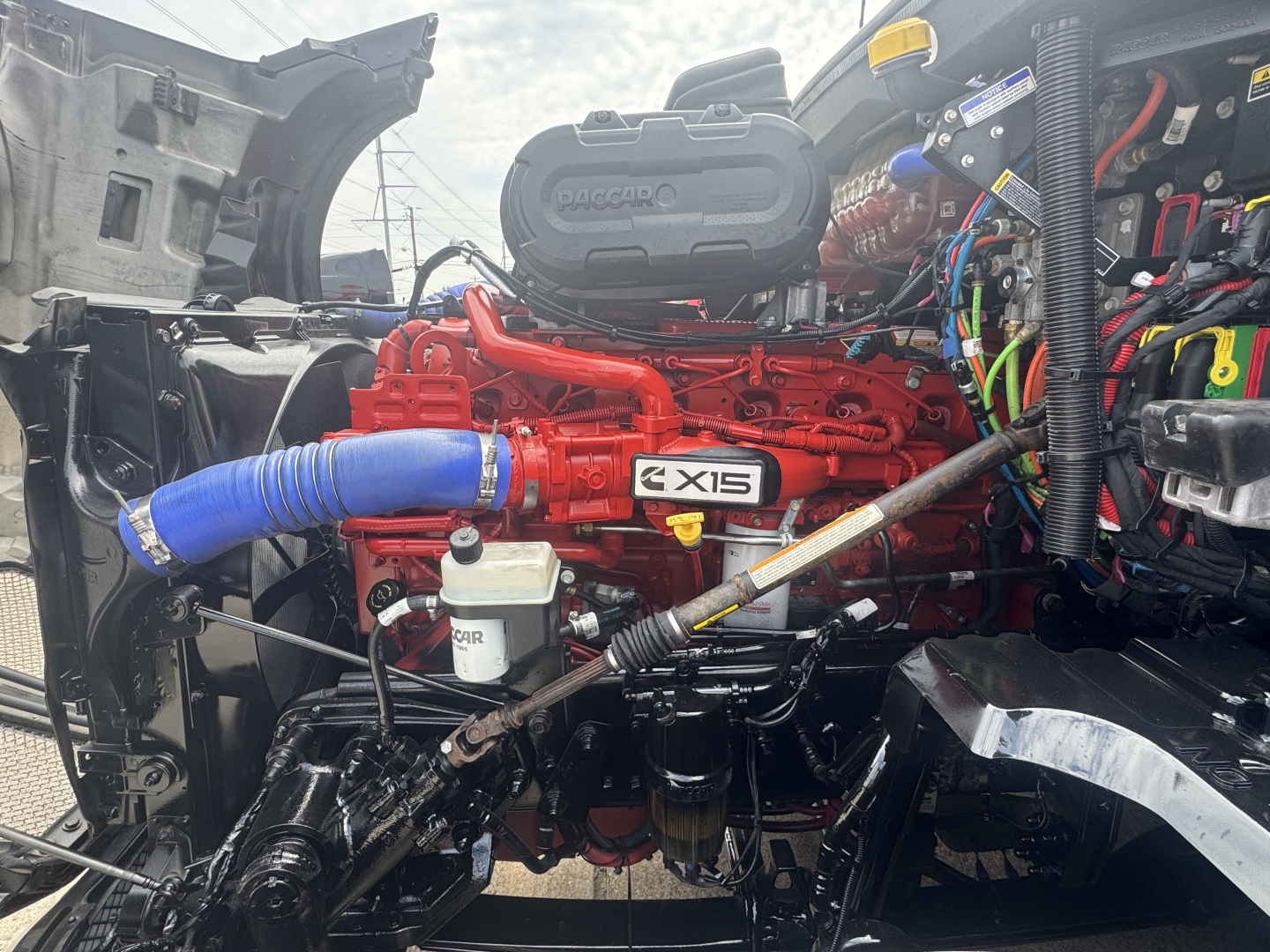 Kenworth T680 engine bay showing 6.8L? red engine with blue intercooler hose, Battery box, wiring harnesses, and various components visible in the bay.