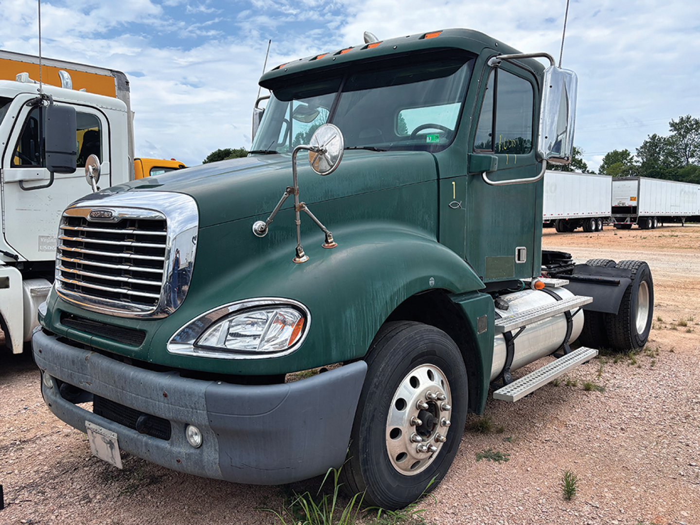 Truck cab: green 2007 Freightliner CL120 Columbia daycab, front-left view showing chrome grille, large round mirrors, dual fuel tanks, steps, and emblem; background includes other trucks and gravel lot.