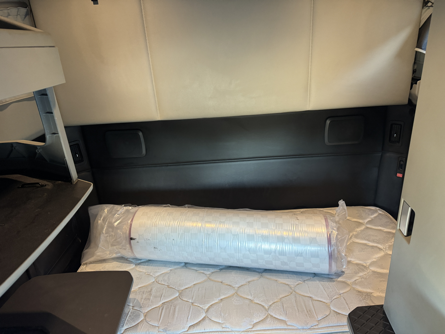 Kenworth T680 sleeper cab interior showing cargo area with a rolled mattress/foam pad in plastic, side storage panels, and beige upholstery.