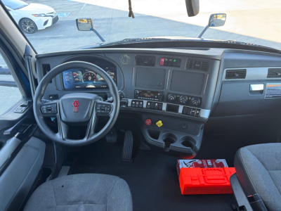 Kenworth T680 truck cab interior showing steering wheel, dashboard, and center console with gauges; passenger seat and red tool box on floor.
