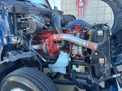 Truck engine bay of a 2024 Kenworth T680 showing red engine, hoses, coolant reservoir, and surrounding components.