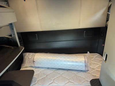 Kenworth T680 sleeper cab interior showing cargo area with a rolled mattress/foam pad in plastic, side storage panels, and beige upholstery.