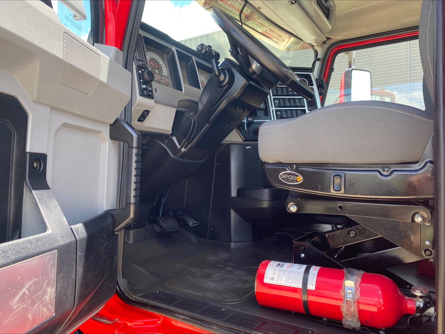 Interior of a 2021 Mack AN64T truck cab showing driver seat, steering wheel, dashboard, pedals, and a red fire extinguisher on the floor.