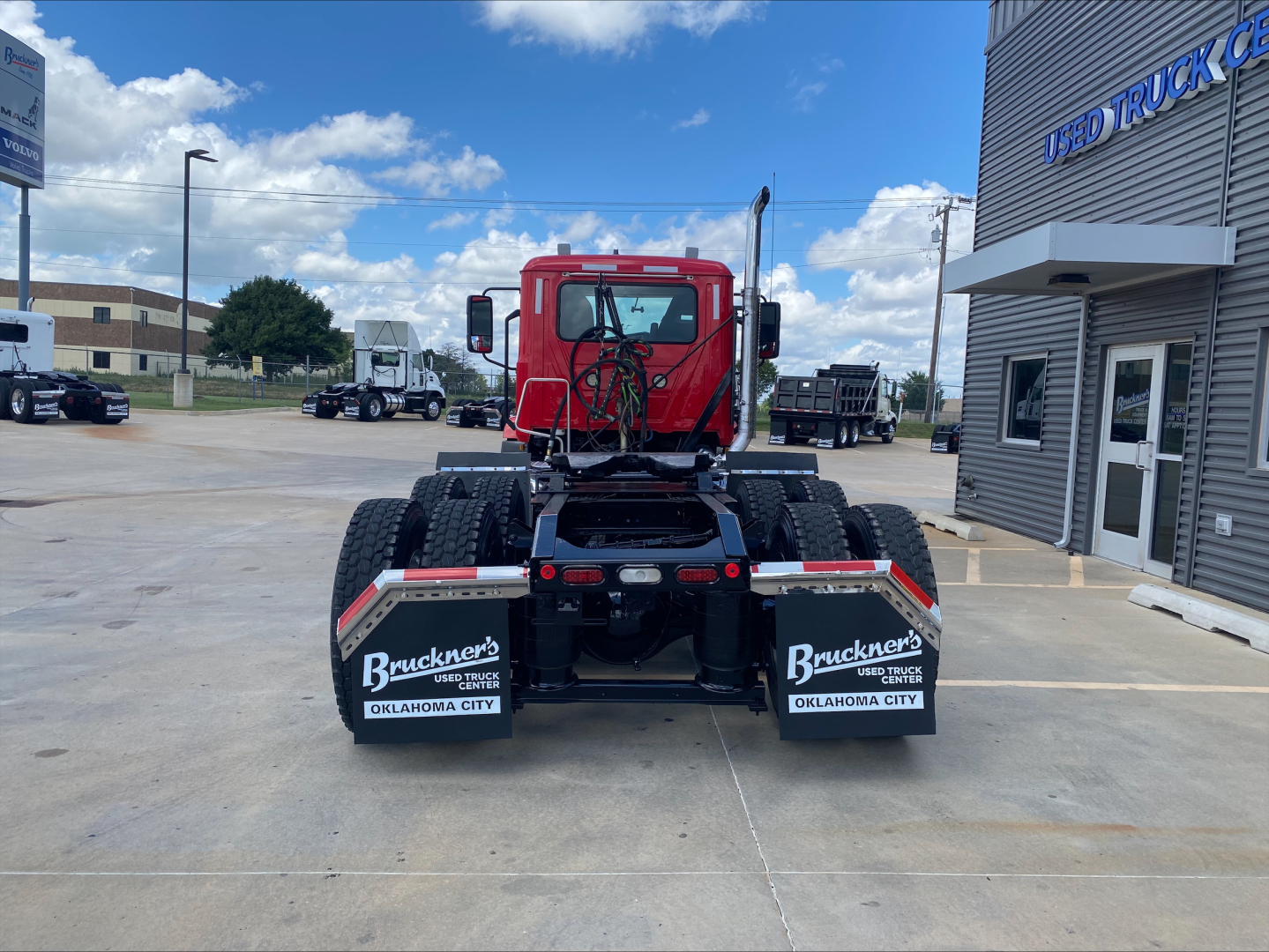 A red Mack AN64T daycab tractor viewed from the rear, showing dual rear axles, mud flaps with Buckner’s OKLAHOMA CITY branding, and a truck dealership building in the background.