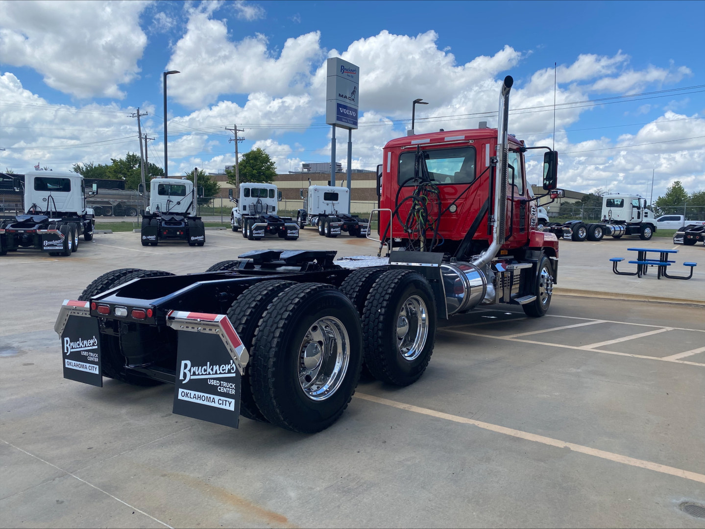 Mack AN64T truck tractor rear and side view showing dual rear axles, red cab, chrome exhaust stacks, and fifth-wheel hitch; dealership lot in background with other trucks.