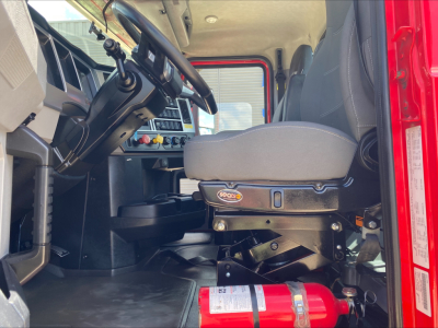 Truck cab interior showing driver seat and dashboard of a 2021 Mack AN64T, with steering wheel, control panel, and a red fire extinguisher mounted near the floor.