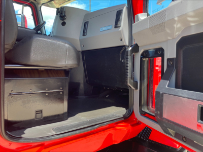 Interior cabin of a red Mack AN64T daycab showing the dashboard and door area, with a storage box under the seat and a black glove compartment.