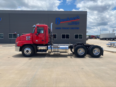 Truck chassis with red cab and dual rear axles in front of a dealership building.