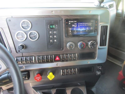 Dashboard of a Western Star 49X truck showing instrument gauges, climate controls, ignition, hazard button, digital radio display, and various switches; visible text includes ‘94.5’, emergency stop red knob, yellow knob, and gear select