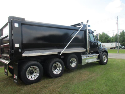 Dump truck side view showing large black dump bed with chrome step rails and dual rear wheels under green grass.