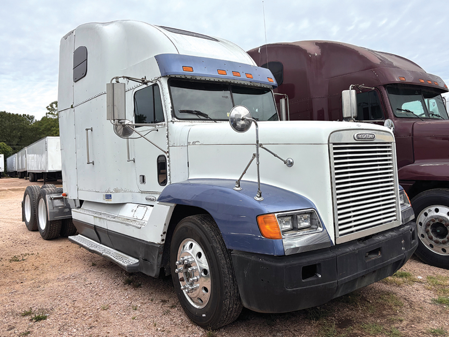 Truck cab showing Freightliner FLD120 sleeper with white cab and blue fender; chrome grille and mirrors; dual rear wheels; adjacent maroon truck in background.