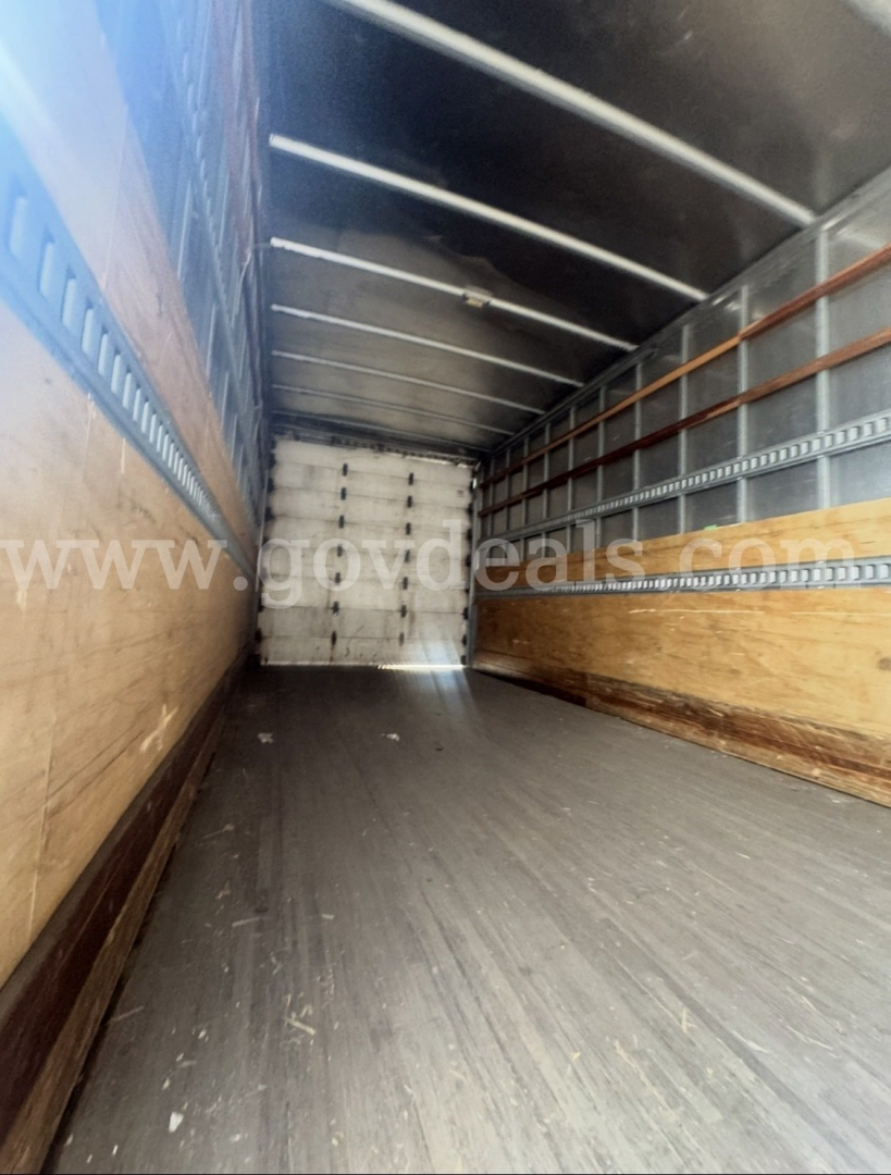 Interior of a truck trailer with wooden side walls and metal railings, empty cargo area seen lengthwise toward the cab, light from open rear end.
