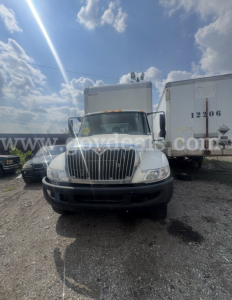 International 4300 box truck front view showing grille, bumper, headlights, and cab with orange roof lights; white body with black bumper; surrounding yard with trailers.