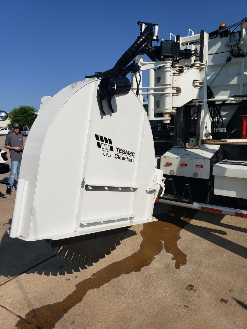 Truck component showing Tesmec Cleanfast attachment with white protective housing and hydraulic crane behind, on a concrete surface.