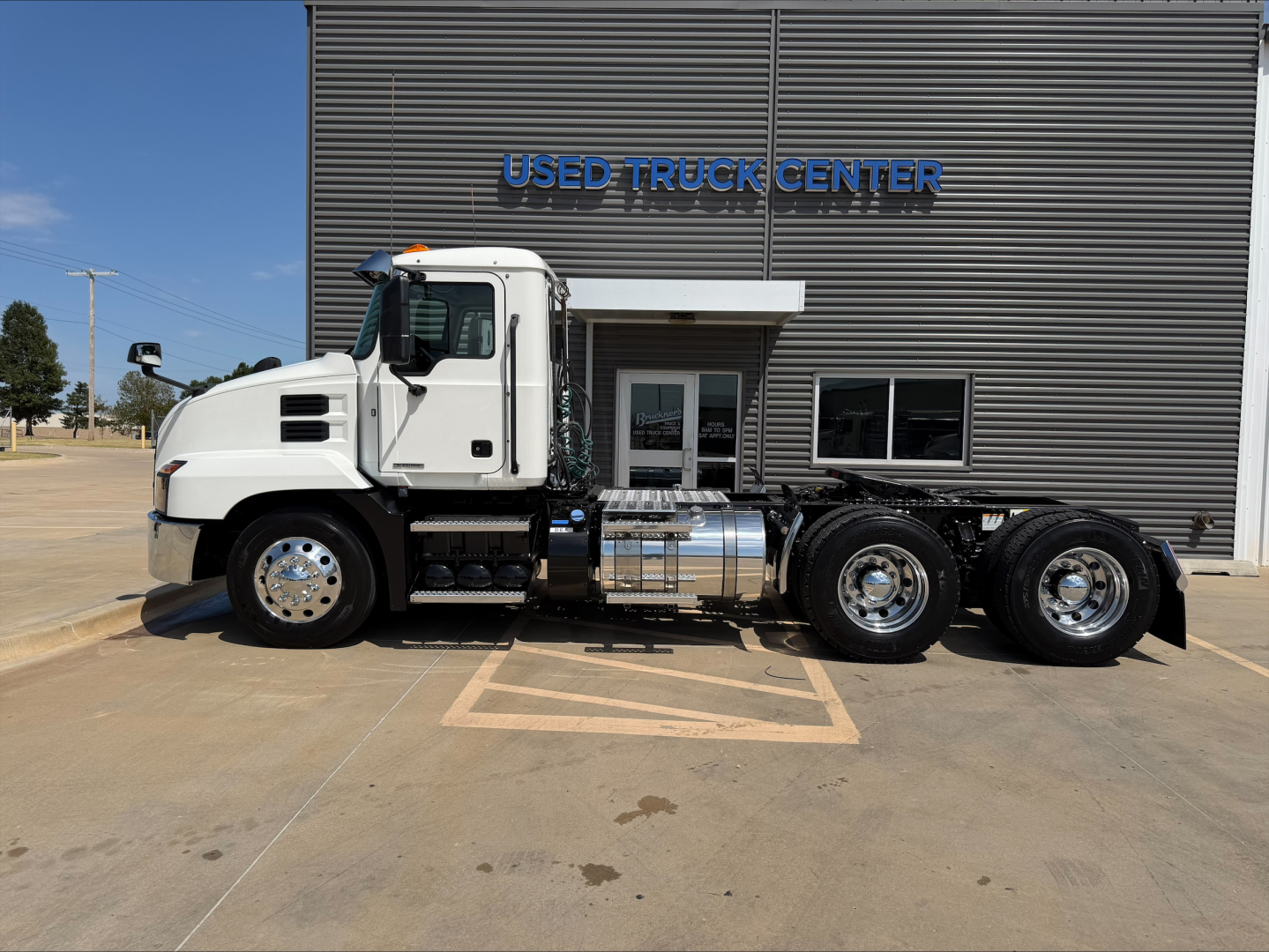 Truck side view showing 2020 Mack ANTHEM conventional daycab with white cab and multiple rear axles in front of a used truck center building.