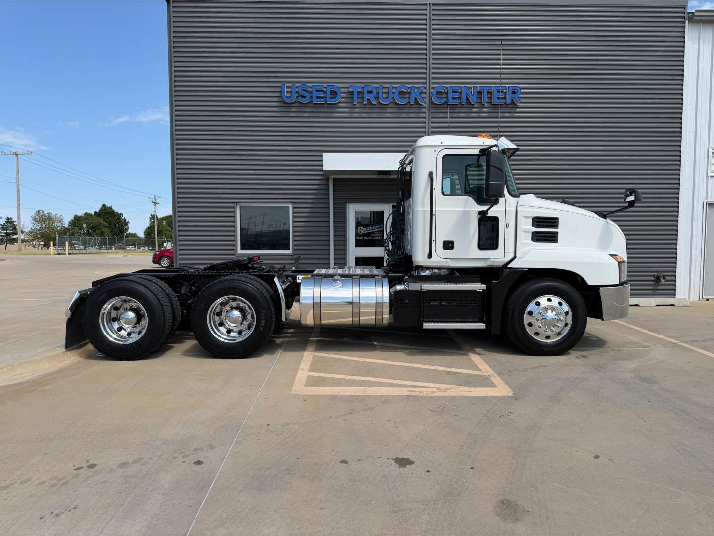 Truck side view of a 2020 Mack ANTHEM conventional daycab in front of a used truck center. Captioned components include dual rear axles, chrome fuel tank, and white cab with black accents.