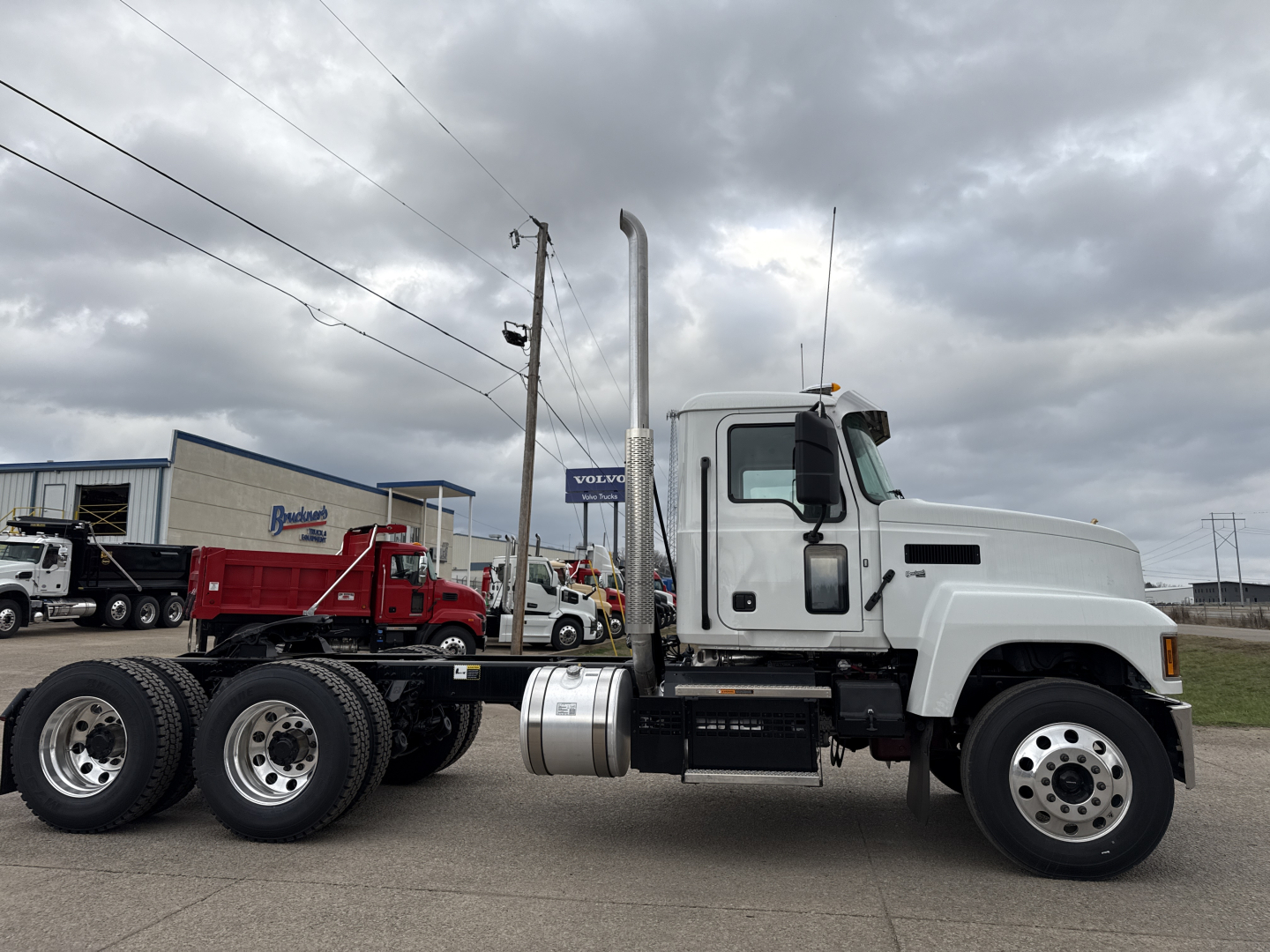 Truck side view: 2026 Mack PI64T conventional daycab with white cab, dual rear axles, fuel tank under cab, vertical exhaust stack, and chrome wheels.