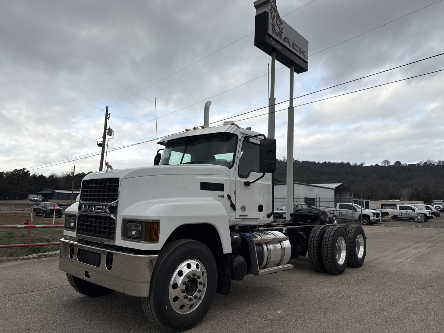 Mack PI64T conventional daycab tractor showing front grille with Mack badge and large headlights; dual rear axles; fuel tanks under cab; parking lot with other trucks in background.