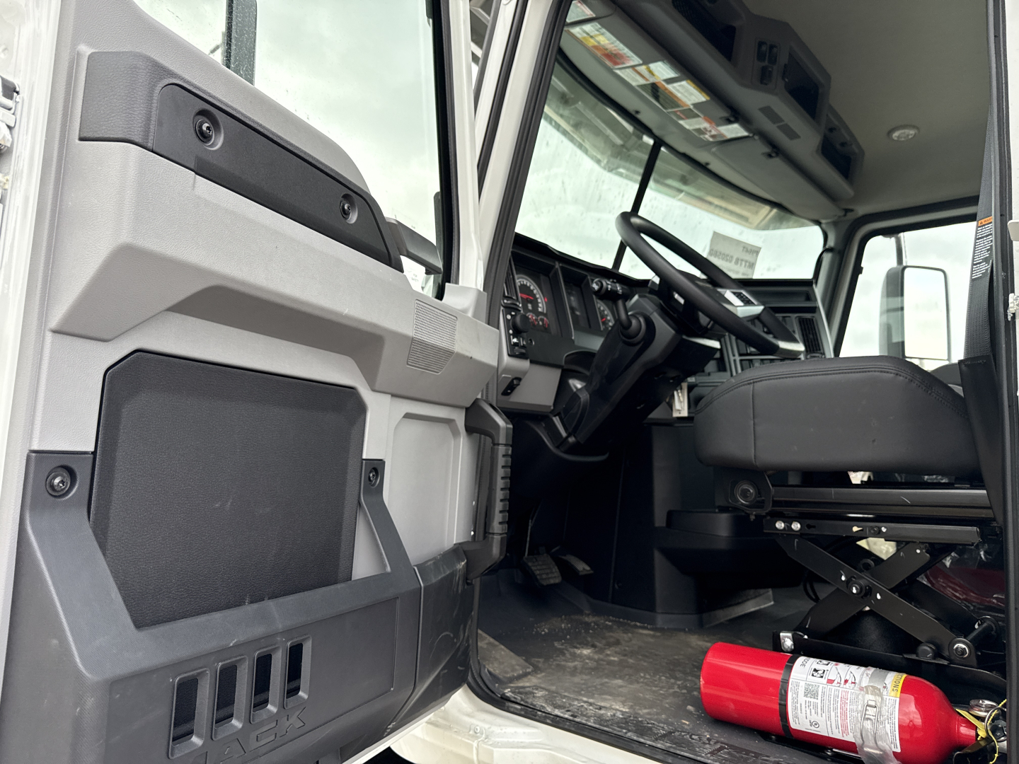 Cab interior of a 2026 Mack PI64T truck showing steering wheel and driver seat, with a red fire extinguisher mounted near the floor and open door.
