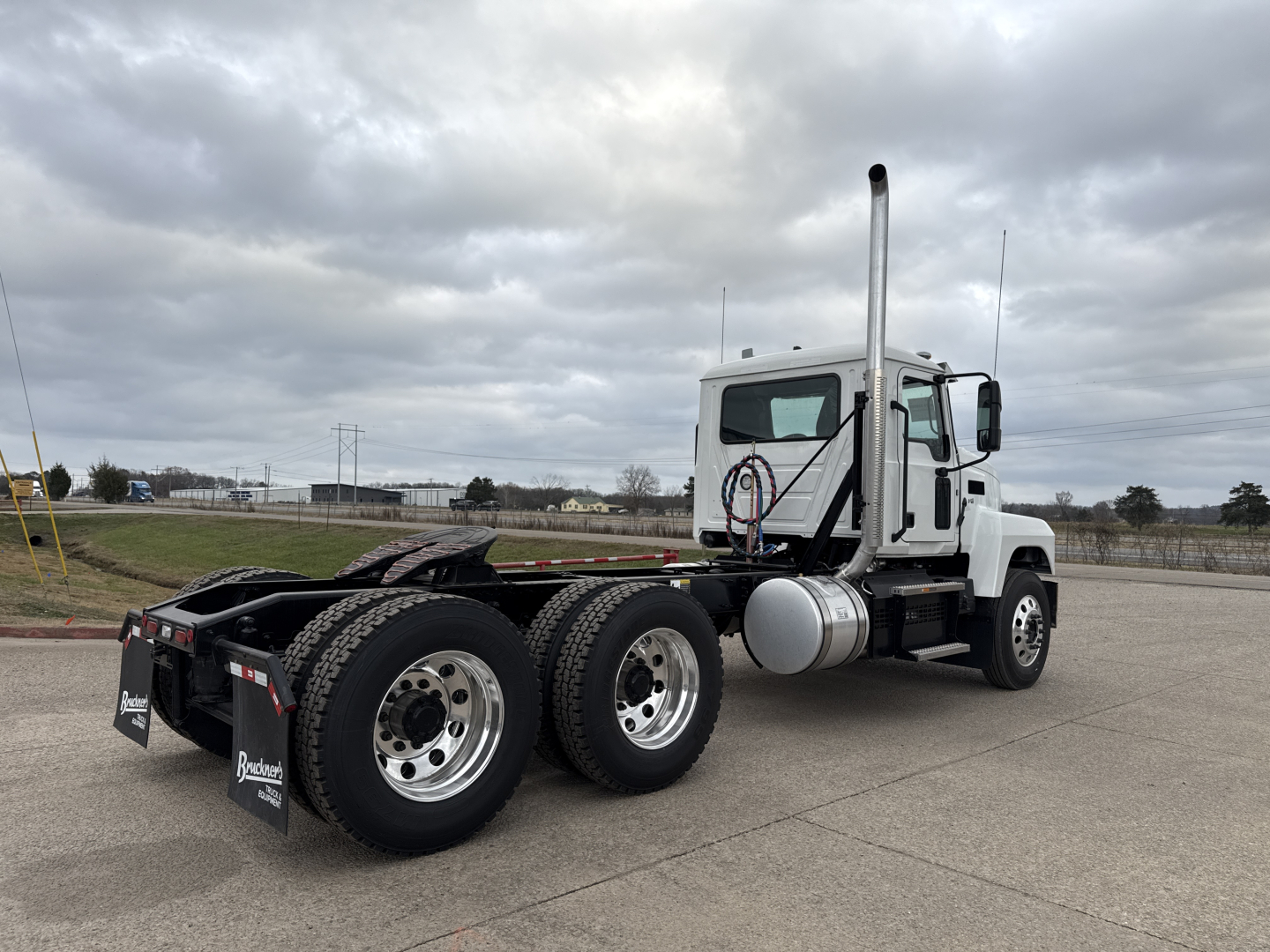 Conventional daycab truck chassis with dual rear axles and vertical exhaust stack, white cab, parked in an open lot. Visible fuel tanks and chrome wheels.