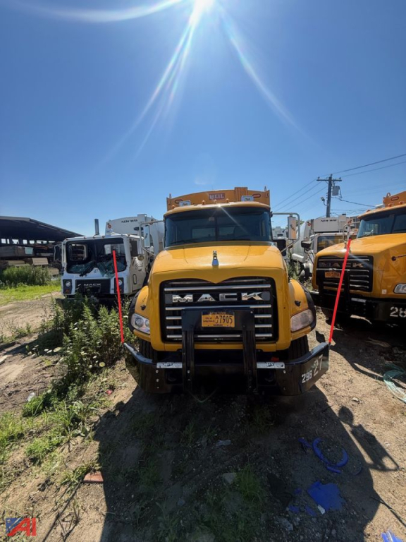 Yellow Mack garbage truck front view with bumper guards and grille, in-service equipment, other trucks in background.