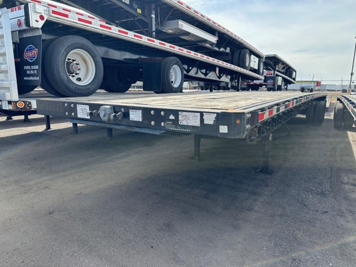 Flatbed trailer with a stacked car carrier on top, rear and side view showing tires and reflective tape. Small stickers and labels visible on the frame near the hitch and side rails.