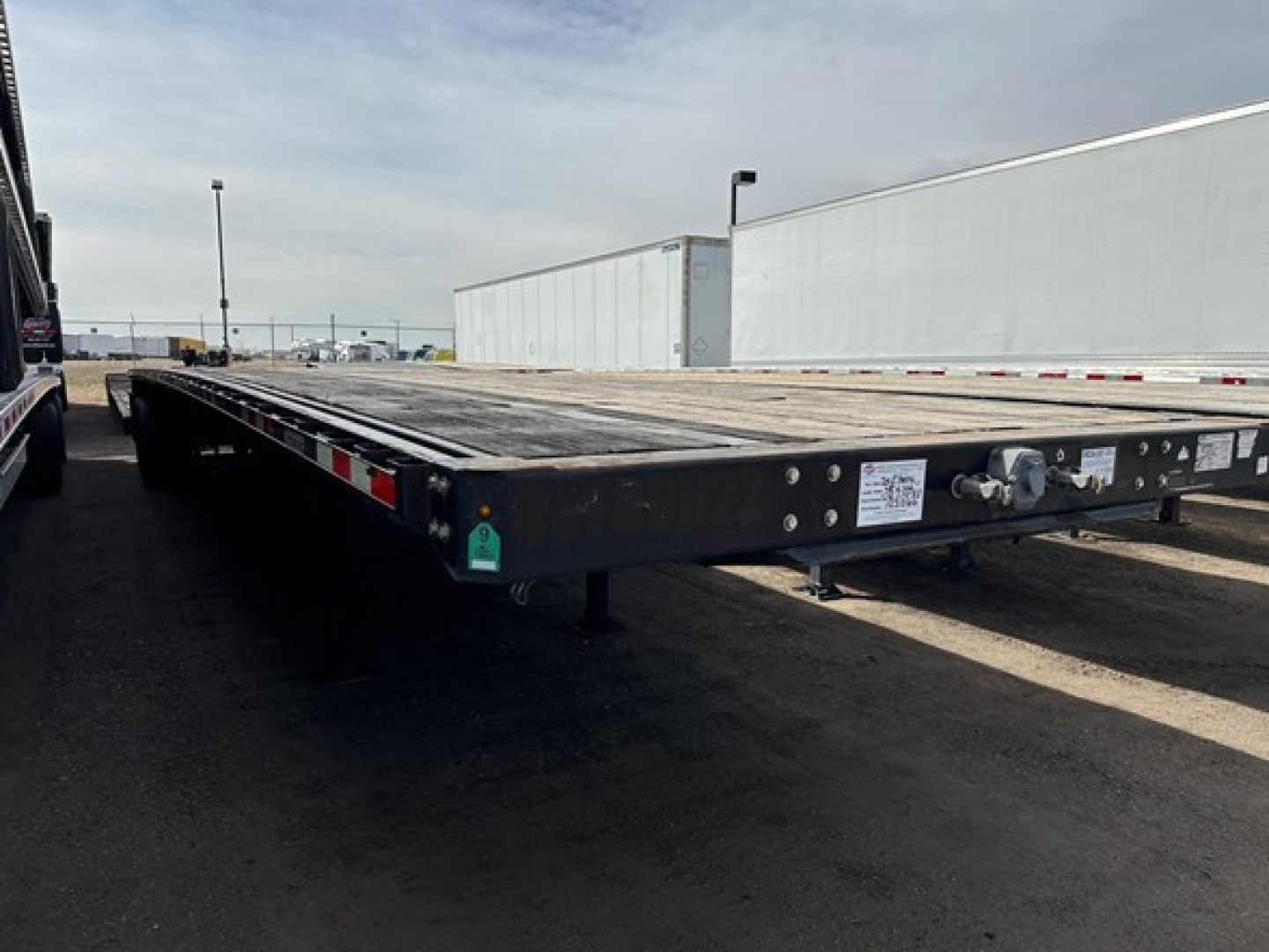 Flatbed trailer showing a 2015 Manac 48' steel flatbed with a low-angle forward view; visible are tie-down points, reflective tape along rails, and label plates near the hitch.