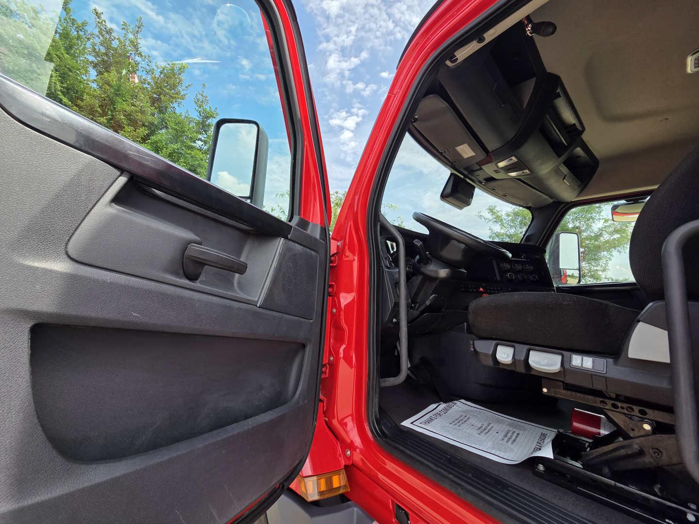 Truck cab interior view from open door showing red exterior, dashboard, driver seat, and floor mats.