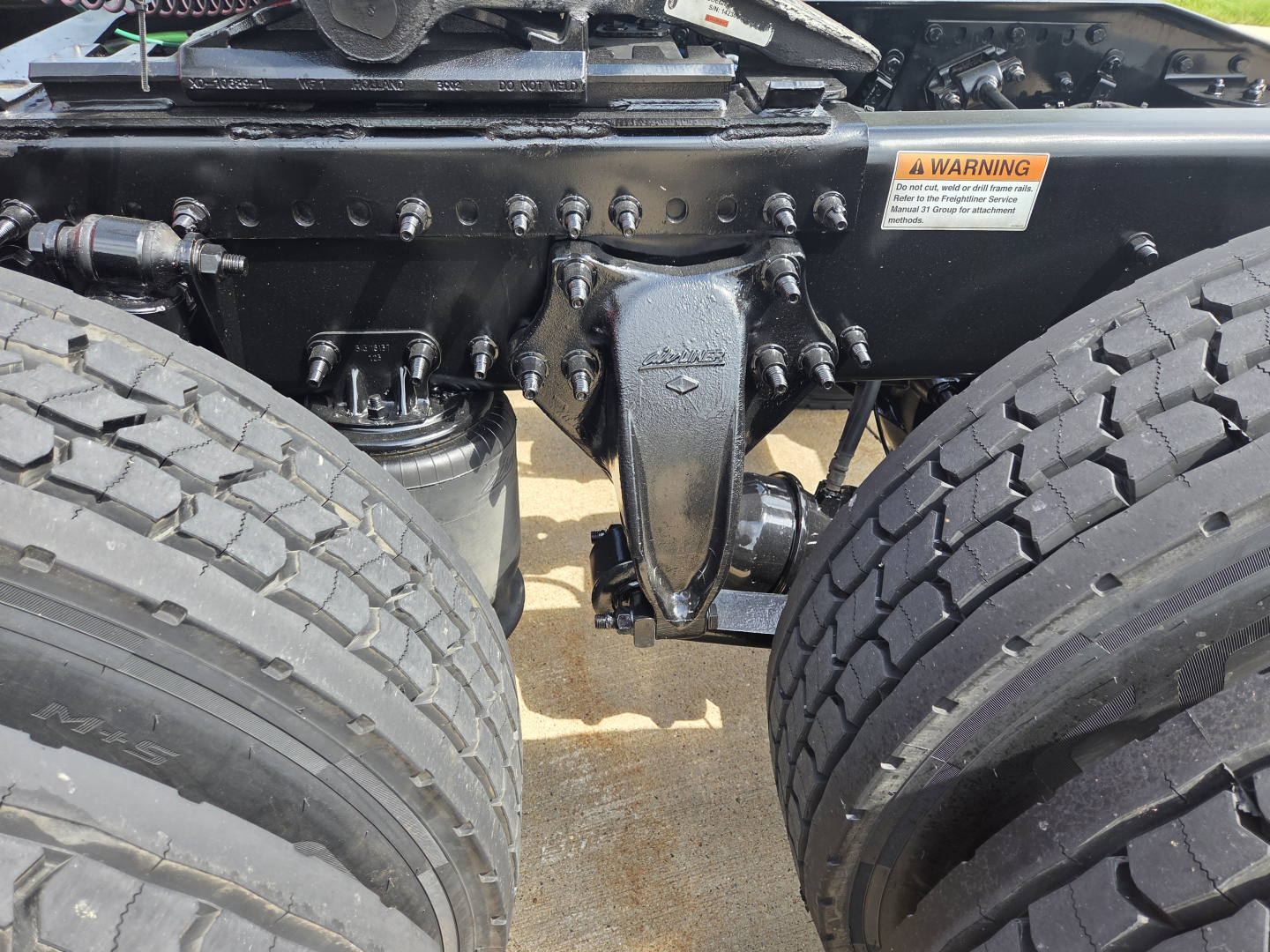 Truck chassis showing suspension and air lines with a warning label; visible components include dual rear axles and a center lightweight frame member.
