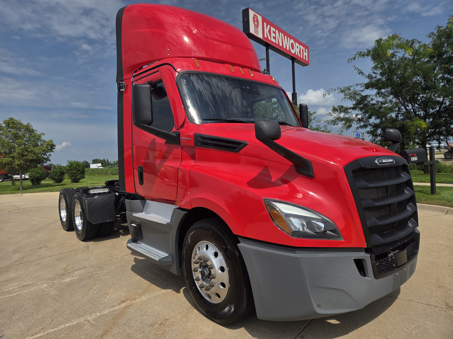 Red Freightliner PT126064S T daycab tractor showing front grille, bumper, dual rear axles, and Kenworth dealership sign in background.