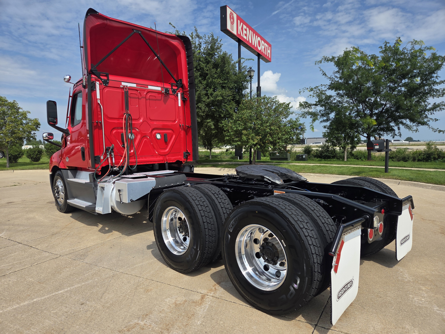 Freightliner PT126064S T daycab truck chassis rear view showing dual rear axles and fifth wheel; red cab with open hatch and Kenworth sign in background.