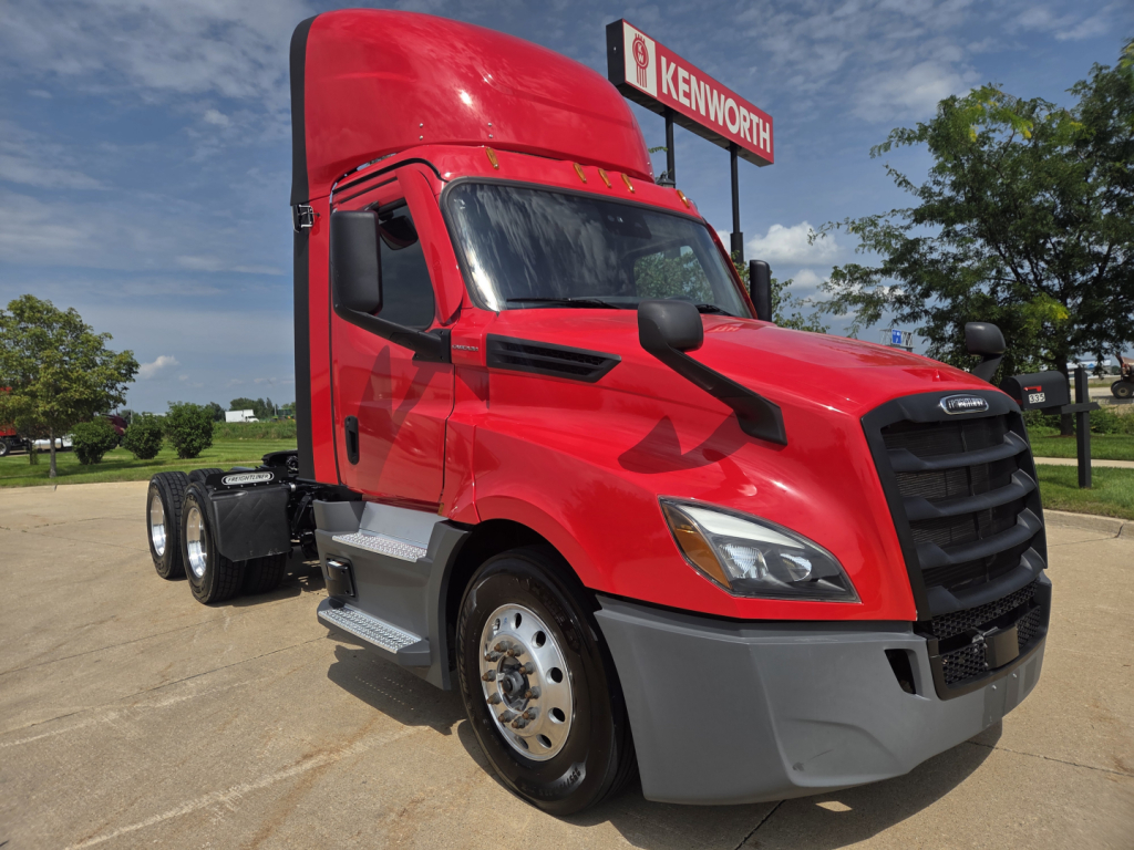 Red Freightliner PT126064S T daycab tractor showing front grille, bumper, dual rear axles, and Kenworth dealership sign in background.