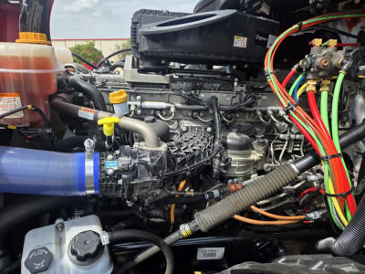 Truck engine bay showing 2024 Freightliner PT126064S T engine with yellow dipstick, blue radiator hose, multiple colored electrical harnesses, and fluid reservoirs.