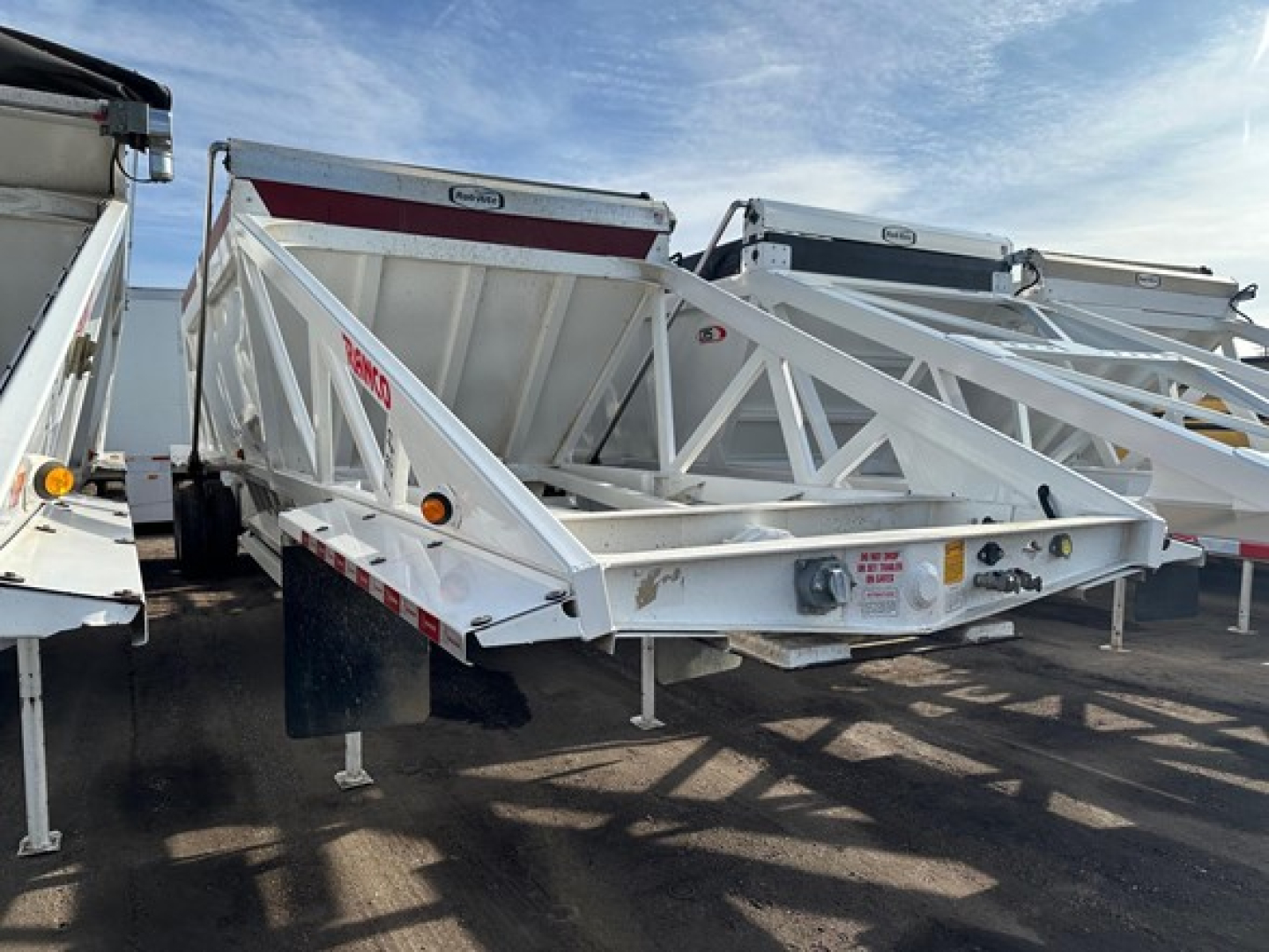 Ranco LW21-40 dump trailer in a lot, white steel with triangular frame and ramp; visible side marker lights and reflection; other trailers in background.