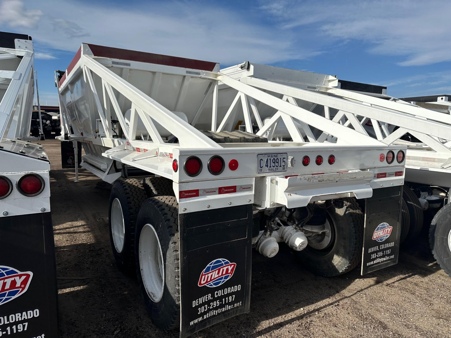 Trailer rear view of a white dump trailer with dual axles and lattice framework, tail lights, mud flaps, and a license plate visible.