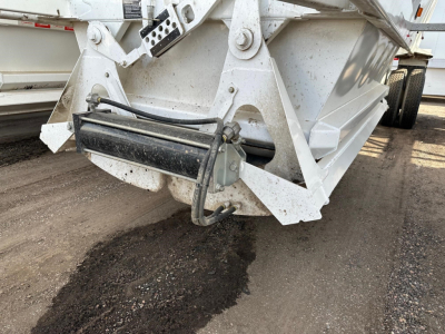 Dump trailer hitch area showing hydraulic hose connections and metal framework. Visible: black hydraulic cylinder/ram at front with hoses, white-painted steel beams, and a portion of a dump bed.