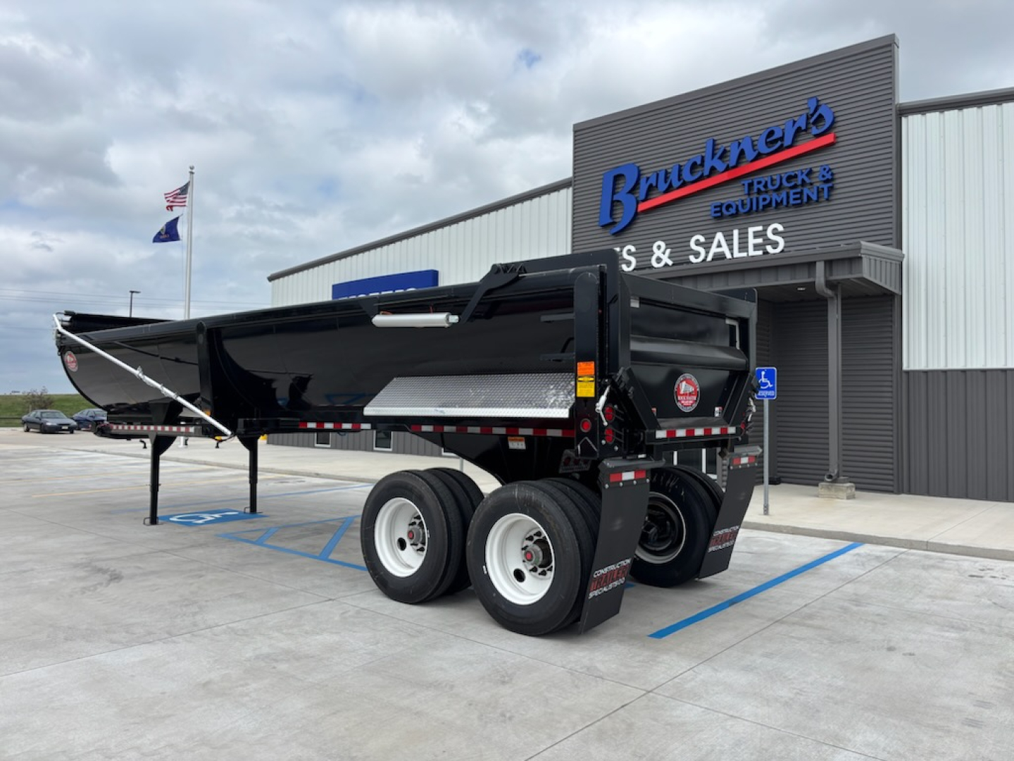 Dump trailer in front of dealership with dual rear axles and black body, side ramp, and Brackner's equipment sign in background.