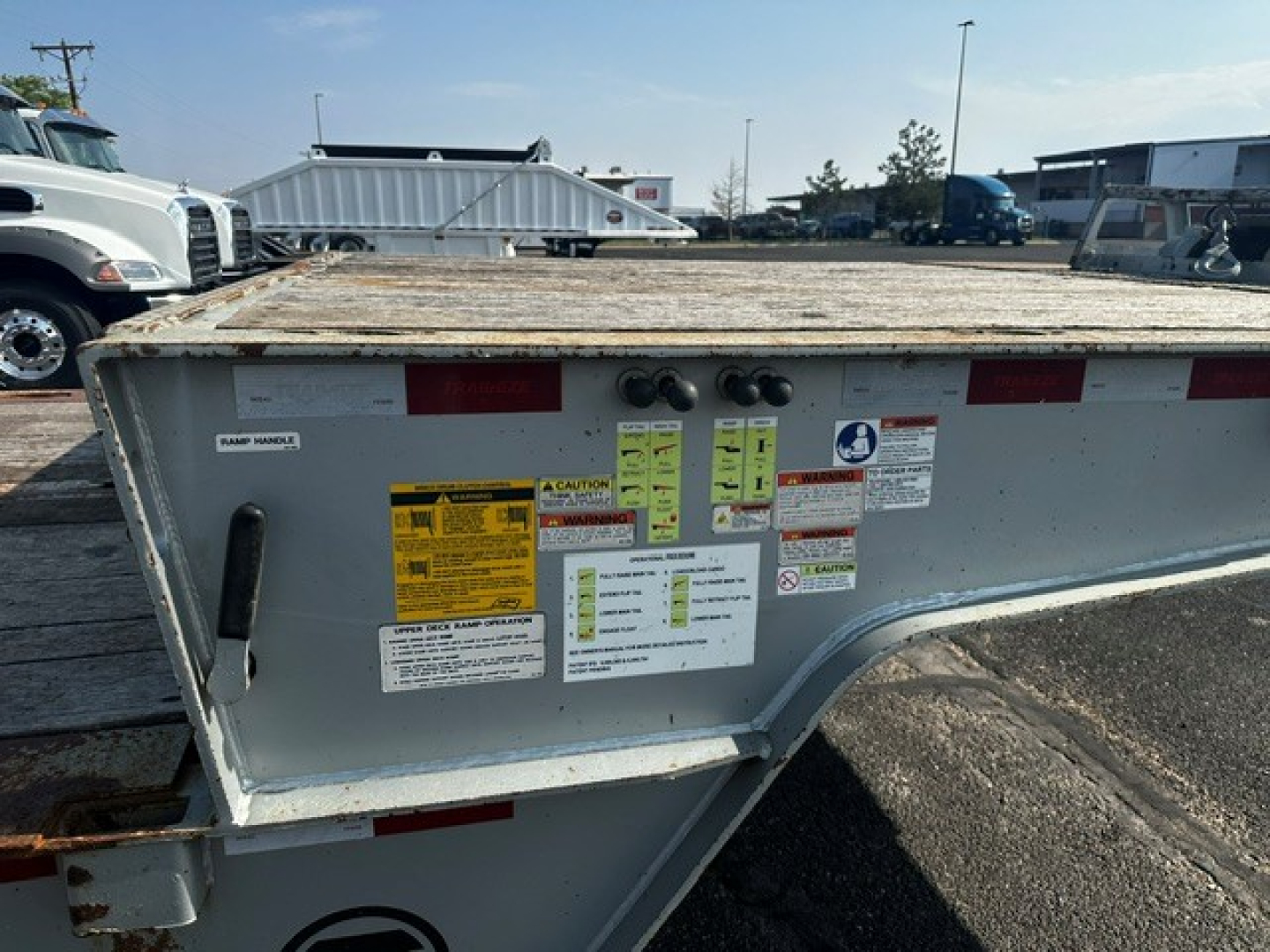 Trailer tailgate with multiple safety and instruction stickers, metal frame, reflective red tape, yellow caution labels, and hydraulic/lighting connections visible.