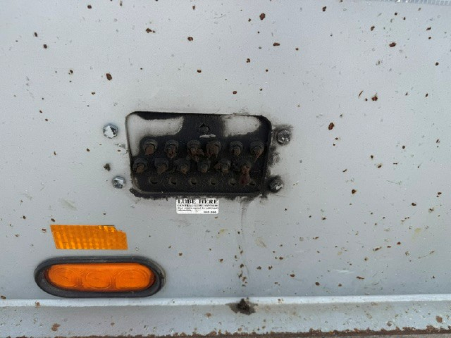 Trailer side showing a damaged or disconnected light and a small identification plate with illegible text; several rust spots on light gray surface.