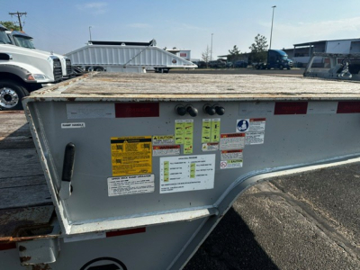 Trailer tailgate with multiple safety and instruction stickers, metal frame, reflective red tape, yellow caution labels, and hydraulic/lighting connections visible.