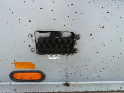 Trailer side showing a damaged or disconnected light and a small identification plate with illegible text; several rust spots on light gray surface.