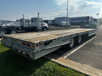 Trailer side and rear view showing a metal deck trailer with dual axles and rear lights, 'TRAIL- EZE' branding, in a parking lot.