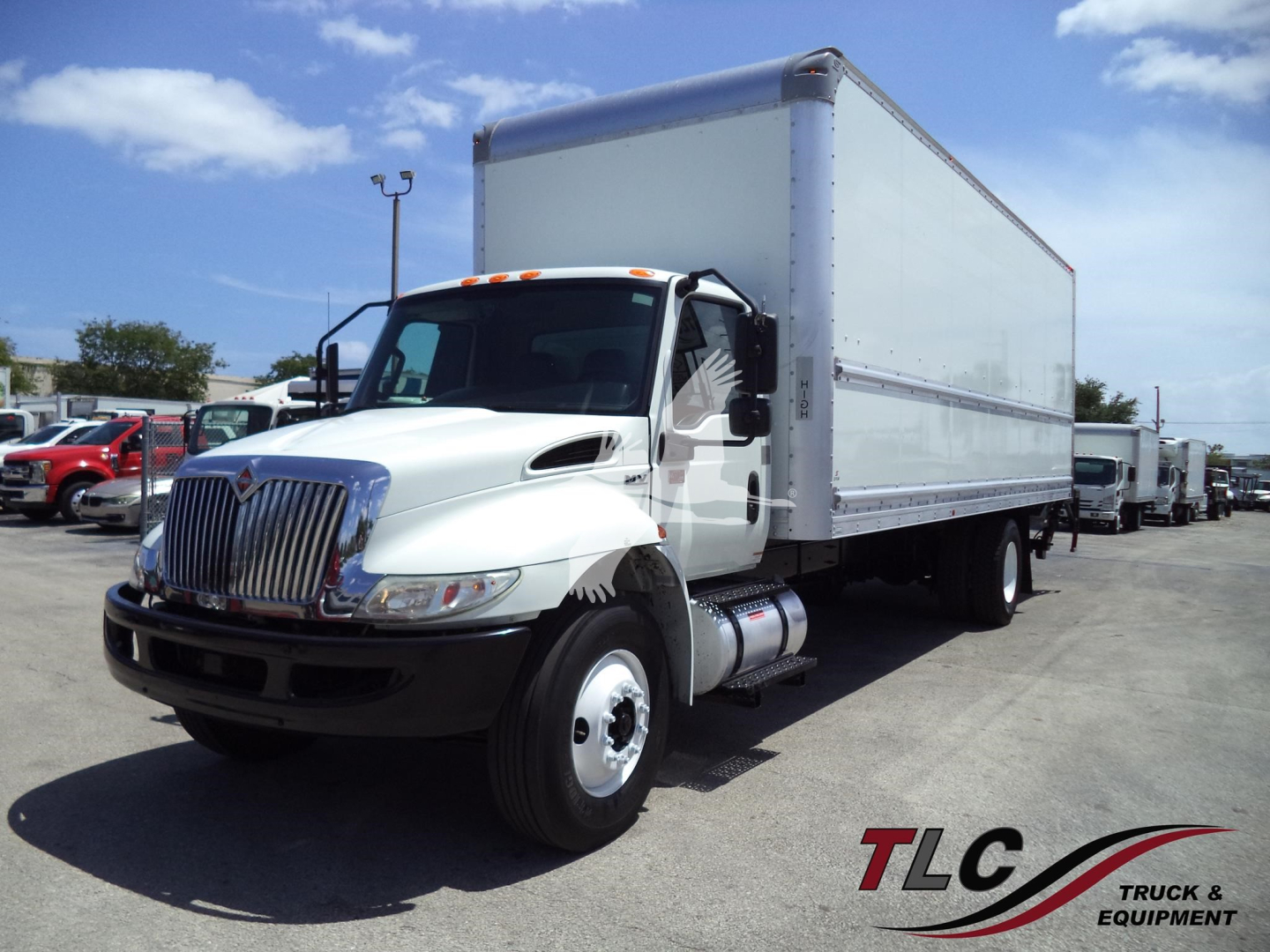 Box truck with white cab and large box body, International logo on front grille, parked in lot with other trucks.